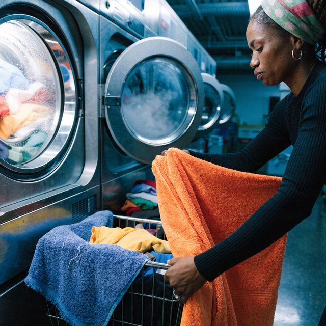 Candid documentary photograph of an adult laundromat attendant sorting warm clothes beside humming machines, cool fluorescent light, 50mm lens with moderate depth of field, loose lint clinging to a blue towel and faint condensation on the glass doors. — Recraft v4