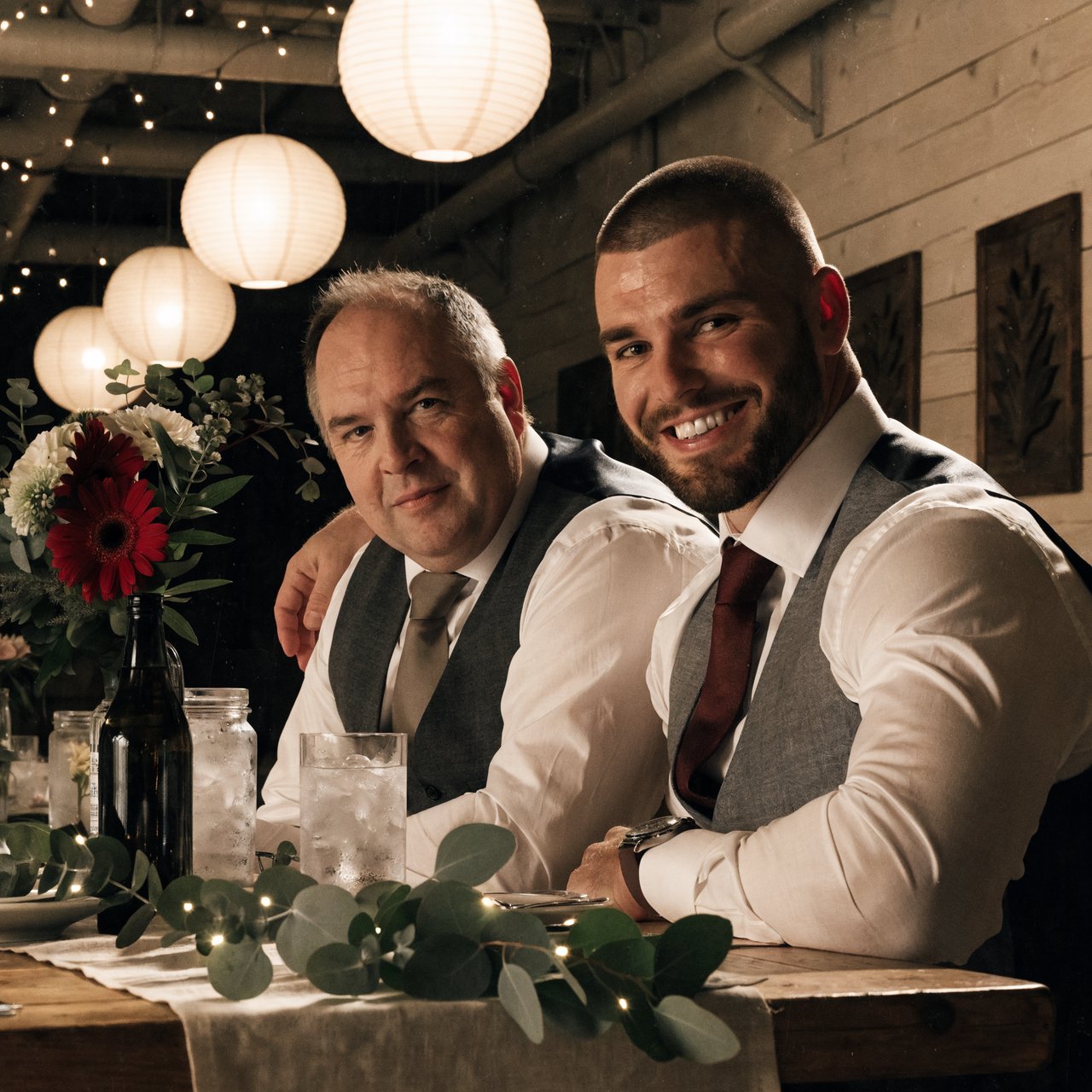(Character1: rugby player body type, tall, brunet male, age 30, buzzcut hair and neatly groomed matching beard). Medium eye-level shot, photorealistic, two men seated at a wooden banquet table: the left middle-aged man wears a white dress shirt, charcoal waistcoat, and taupe tie, with his right arm on the table; the right man, Character1, smiles directly at the camera, wearing a white dress shirt, grey waistcoat, maroon tie, and a watch on his left wrist, with his left arm draped over the first man's shoulder. In the foreground: a table runner with eucalyptus sprigs, small fairy lights, a bottle holding flowers, a glass jar, and a water glass. Background: an exposed light horizontal wood plank wall with two decorative leaf panels, an exposed industrial ceiling with hanging paper lanterns, and fairy string lights. Warm, soft ambient overhead event lighting, natural indoor venue atmosphere, shallow depth of field. Art style: dramatic painterly realism with realistic skin texture, strong chiaroscuro, slightly gritty textured brushwork, fantasy illustration mood. — Recraft v4 Pro