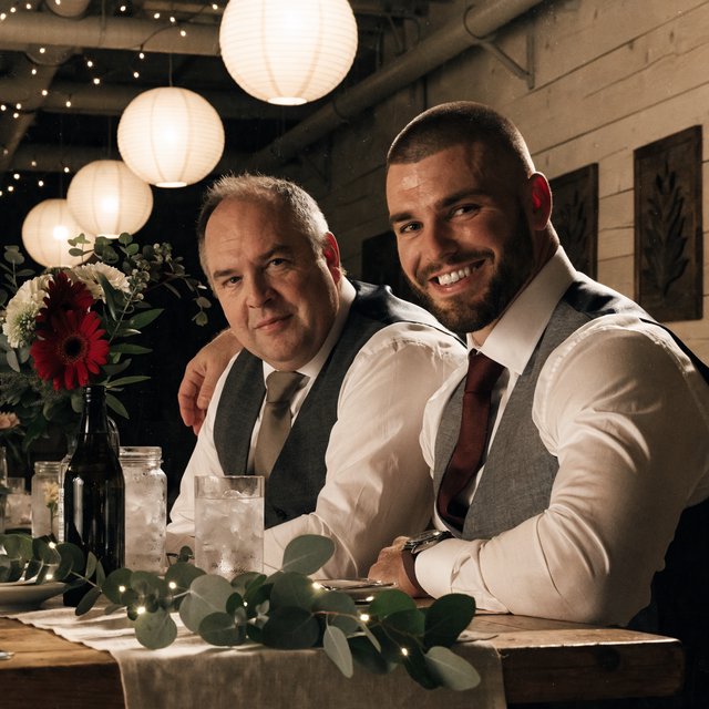 (Character1: rugby player body type, tall, brunet male, age 30, buzzcut hair and neatly groomed matching beard). Medium eye-level shot, photorealistic, two men seated at a wooden banquet table: the left middle-aged man wears a white dress shirt, charcoal waistcoat, and taupe tie, with his right arm on the table; the right man, Character1, smiles directly at the camera, wearing a white dress shirt, grey waistcoat, maroon tie, and a watch on his left wrist, with his left arm draped over the first man's shoulder. In the foreground: a table runner with eucalyptus sprigs, small fairy lights, a bottle holding flowers, a glass jar, and a water glass. Background: an exposed light horizontal wood plank wall with two decorative leaf panels, an exposed industrial ceiling with hanging paper lanterns, and fairy string lights. Warm, soft ambient overhead event lighting, natural indoor venue atmosphere, shallow depth of field. Art style: dramatic painterly realism with realistic skin texture, strong chiaroscuro, slightly gritty textured brushwork, fantasy illustration mood. — Recraft v4 Pro