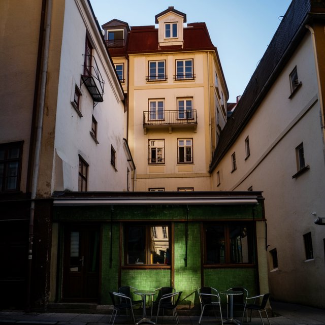 A tiny corner café in a narrow European street, green tiled facade, two bistro tables, morning light, no people. — Krea 2 Large
