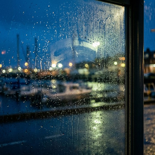 Raindrops crawl down a bus shelter pane, warping blurred harbor lights beyond. A handheld evening street snapshot in cool sodium glow, shot at 50mm with shallow depth of field, catches tiny water trails and a faint wipe mark. — Nano Banana 2.1
