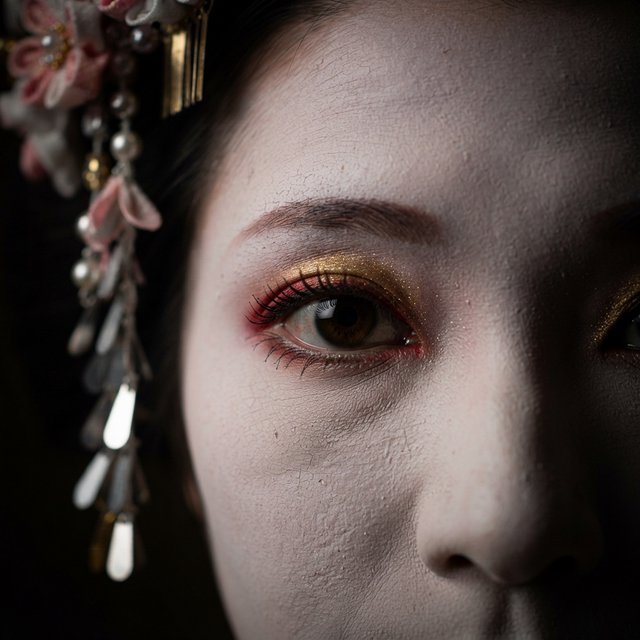 Extreme close-up macro photography of a Geisha's face. The focus is on her eyes, revealing intense emotion and hyper-realistic detail. Her skin is covered in traditional white 'Oshiroi' makeup, showing subtle, realistic cracking and skin texture. Dramatic Chiaroscuro lighting with deep shadows and a single soft light source highlighting the shimmer in her golden eyeshadow and the wetness of her eyes. Ornate kanzashi (hair ornaments) visible in the blurred periphery with intricate silk flowers and dangling silver petals. Cinematic composition, shot on 100mm macro lens, incredibly detailed eyelashes and iris patterns. — Model 16