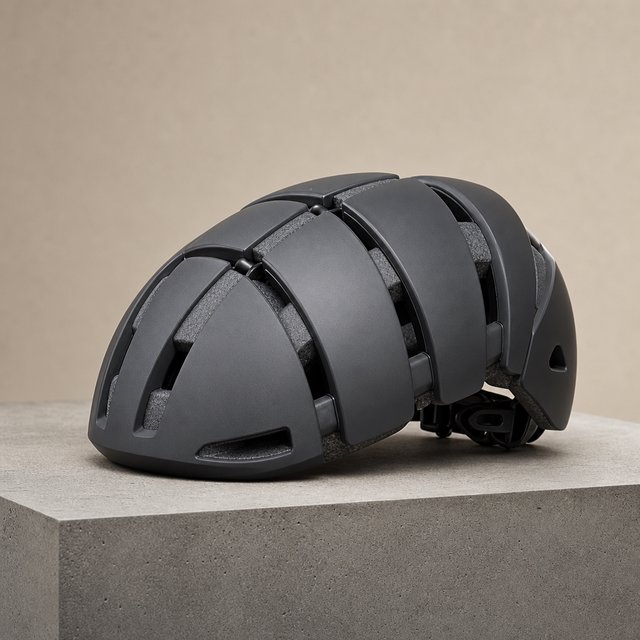 Commercial product photograph of an unbranded folding bicycle helmet made from matte charcoal polymer with soft gray padding, resting on a honed concrete plinth against a muted sand background, illuminated by a broad overhead softbox and gentle rim light. — GPT Image 2