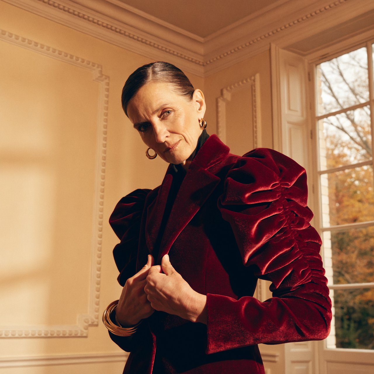 Fashion portrait of an adult woman in her early 50s wearing a sculptural burgundy velvet jacket, her hands lightly gathering its lapel. A wry, composed expression meets the camera against a warm ivory plaster backdrop, lit by soft late-afternoon window glow. — Recraft v4 Pro