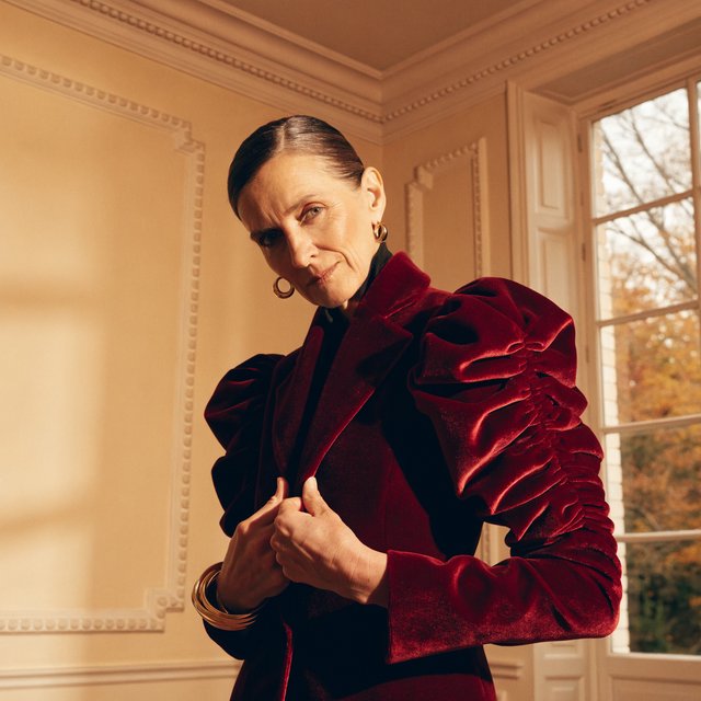 Fashion portrait of an adult woman in her early 50s wearing a sculptural burgundy velvet jacket, her hands lightly gathering its lapel. A wry, composed expression meets the camera against a warm ivory plaster backdrop, lit by soft late-afternoon window glow. — Recraft v4 Pro