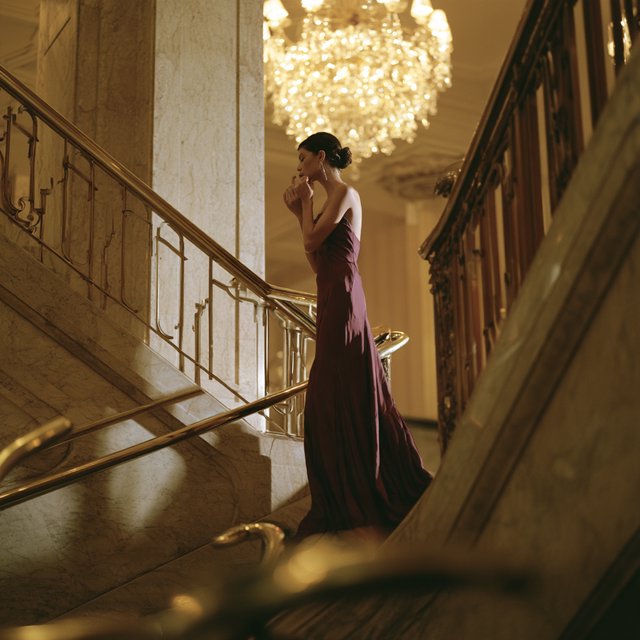 At the landing of a marble staircase, an adult woman in a plum evening gown pauses to fasten an earring, caught in warm chandelier light; polished brass rails and soft shadows lend the scene the poise of a fashion magazine. — Midjourney V8.2