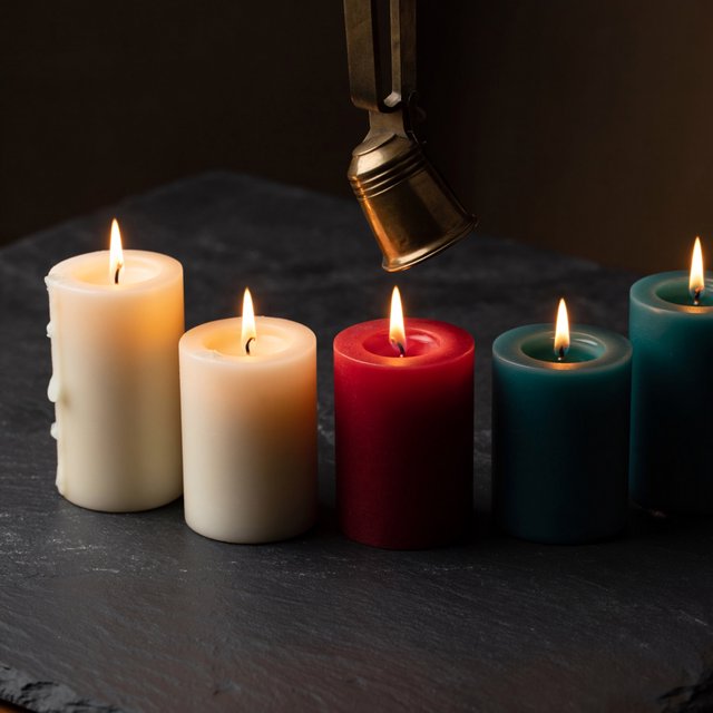 Exactly five candles stand on a dark slate tabletop: two ivory candles on the left, one red candle in the center, and two teal candles on the right. A brass snuffer sits above the red candle, nothing else. — ImagineArt 2