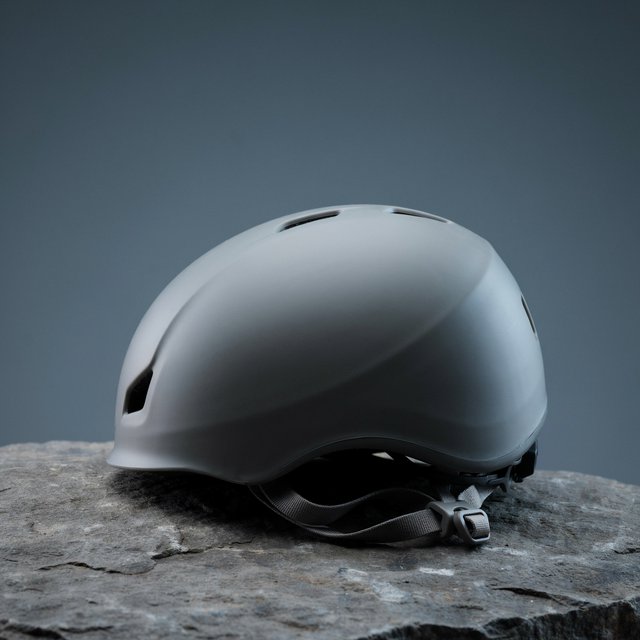 Against a muted slate backdrop, an unbranded climbing helmet with a matte recycled-polymer shell and woven nylon straps rests on rough granite; broad diffused key light and a cool rim light shape this commercial product image, with no lettering. — Ideogram v4