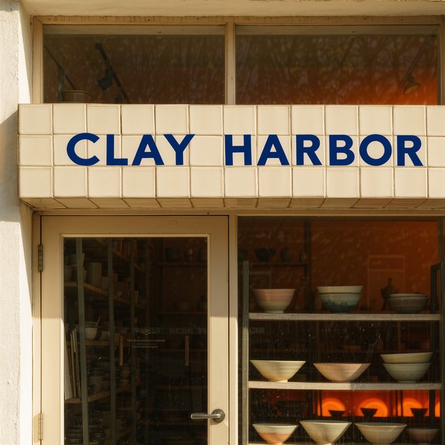 A sunlit ceramicist's studio storefront displays glazed bowls behind dusty glass doors; "CLAY HARBOR" is printed in cobalt-blue block letters across a cream tile transom above the entrance, with kiln warmth glowing inside. — Kling v3 Omni