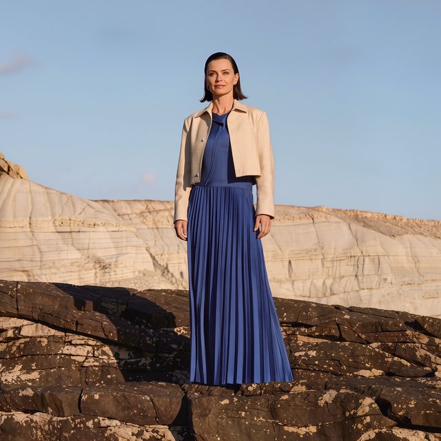 Across a basalt terrace, an adult woman in her early forties poses in a full-length fashion photograph, wearing a pleated indigo silk dress and cropped linen jacket, her composed half-smile lit by hard afternoon sunlight from one side against a pale limestone ridge. — Kling v3