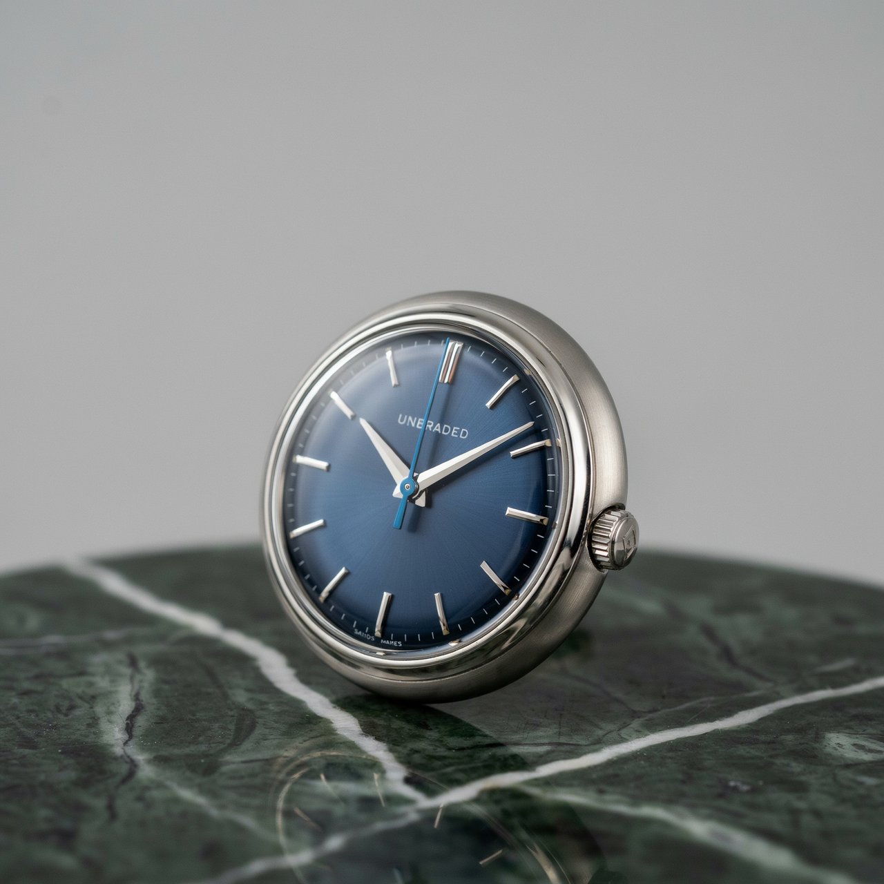 Commercial product photograph of an unbranded mechanical wristwatch with a brushed titanium case and deep blue enamel dial, resting on dark green marble against a soft gray background, illuminated by a large diffused softbox and narrow edge light. — Ideogram v4