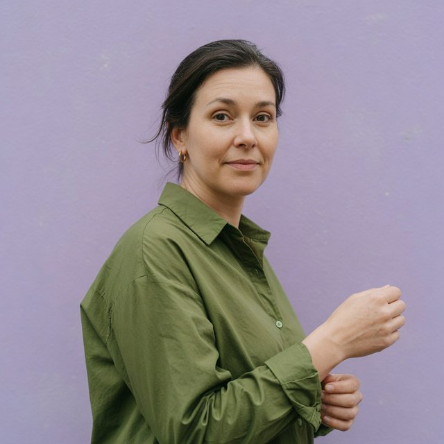 Portrait photograph of an adult woman in her late 30s wearing a moss-green cotton poplin shirt, fingers lightly turning its rolled cuff. Her calm, observant expression faces the camera against a chalky lavender backdrop, lit by soft diffused skylight. — Grok Imagine Image Quality