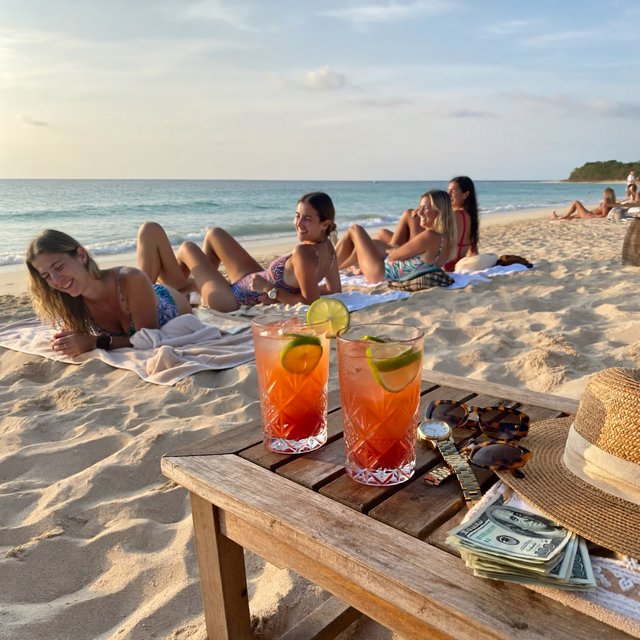 A realistic scene of women in one-piece swimsuits on the beach, with a peaceful vibe, orange and red drinks nearby, high-quality items nearby, a bit of money also nearby, and many angles. — ImagineArt 2