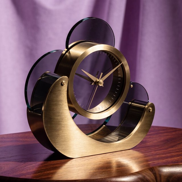 Commercial product photograph of an unbranded sculptural table clock made from satin brass and smoked glass, resting on a dark walnut surface against a muted lavender background, illuminated by a large diffused softbox from the upper left and a subtle rear rim light. — Recraft v4 Pro
