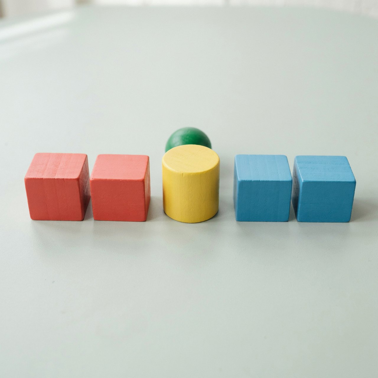 Exactly five wooden toy blocks sit on a pale gray table: two red cubes on the left, one yellow cylinder in the center, and two blue cubes on the right. A small green ball sits behind the cylinder, nothing else. — Ideogram v4.5