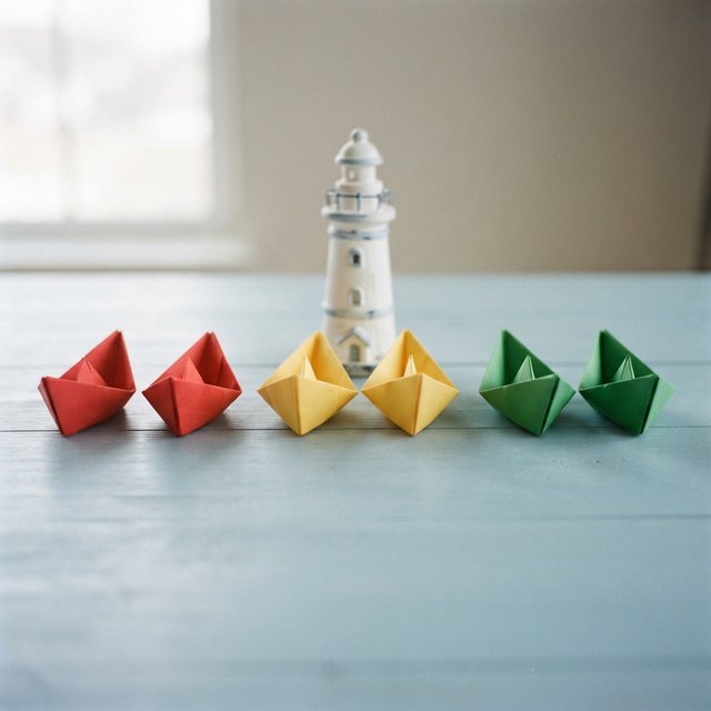 Exactly six folded paper boats sit on a pale blue tabletop: two red boats on the left, two yellow boats in the center, and two green boats on the right. A small white lighthouse figurine stands behind the center pair, nothing else. — Nano Banana 2 Lite