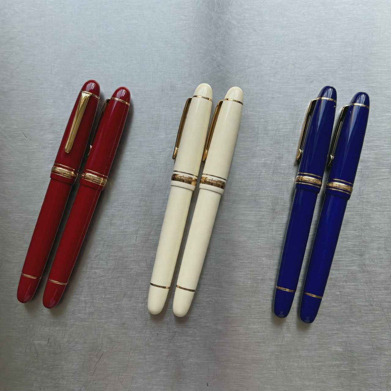 Across a brushed steel counter, exactly six fountain pens rest in three groups: two crimson pens on the left, two ivory pens in the center, and two cobalt pens on the right; nothing else. — Muse Image 1.0
