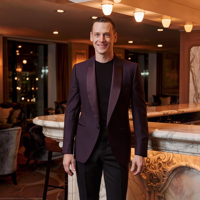 Editorial glamour photograph of an adult man in a midnight-plum dinner jacket and black trousers, standing naturally beside a sculptural marble bar in a quiet hotel lounge, flattering amber light, easy smile, polished editorial elegance. — Kling v3