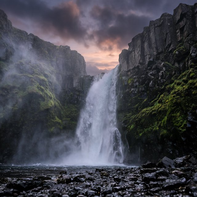 After a passing squall, a waterfall drops through a narrow basalt gorge at dusk, its white plume breaking against dark ledges; rain-beaded moss and sharp foreground stones lead toward cliffs fading behind drifting spray. — Krea 2 Large
