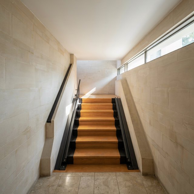 Beneath a pale limestone stairwell, a restrained mid-century modern interior frames a straight oak stair with blackened-steel stringers and limestone landings; afternoon clerestory light, viewed from the upper landing, reveals credible connections and aligned geometry. — Ideogram v4.5