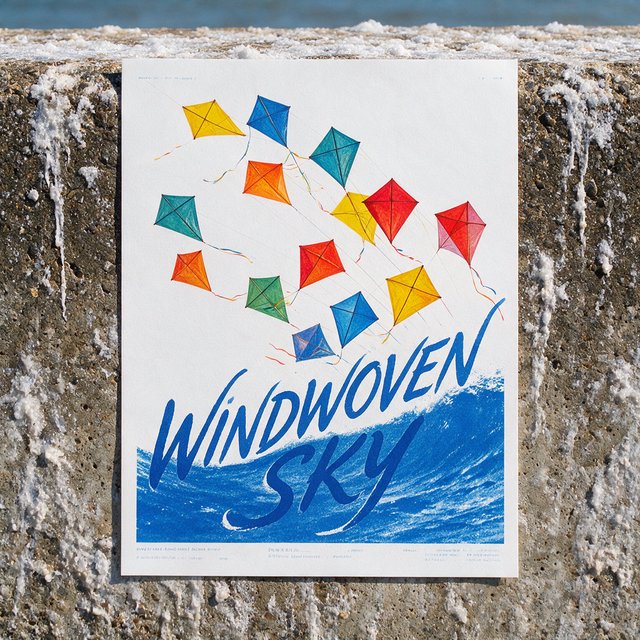 On a salt-stained seawall, a printed poster for a kite-making exhibition shows bright diamond kites sweeping above blue waves; “WINDWOVEN SKY” is screen-printed in sweeping cobalt script, with no other legible writing. — Model 20