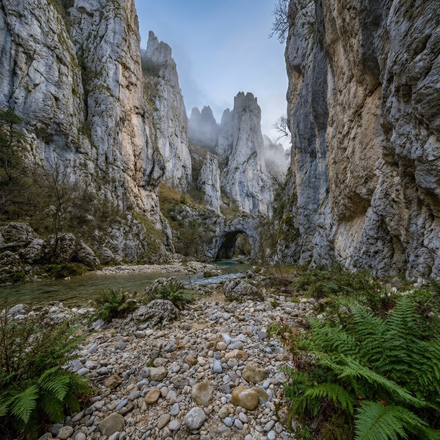 A limestone karst valley at blue hour after a light shower, towering pale cliffs rising above a clear river, low clouds snagging on pinnacles, slick ferns and rounded pebbles leading toward a distant stone arch. — Qwen Image 3.0 Pro