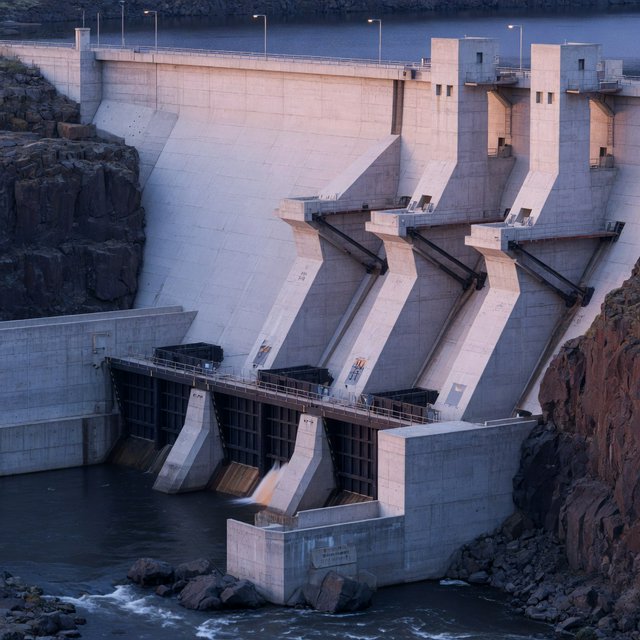 A broad hydroelectric dam in contemporary Brutalist engineering style spans a rocky gorge, its pale concrete face anchored by dark basalt abutments and steel gates; cool dawn light, viewed from downstream, with convincing load-bearing geometry. — Model 6
