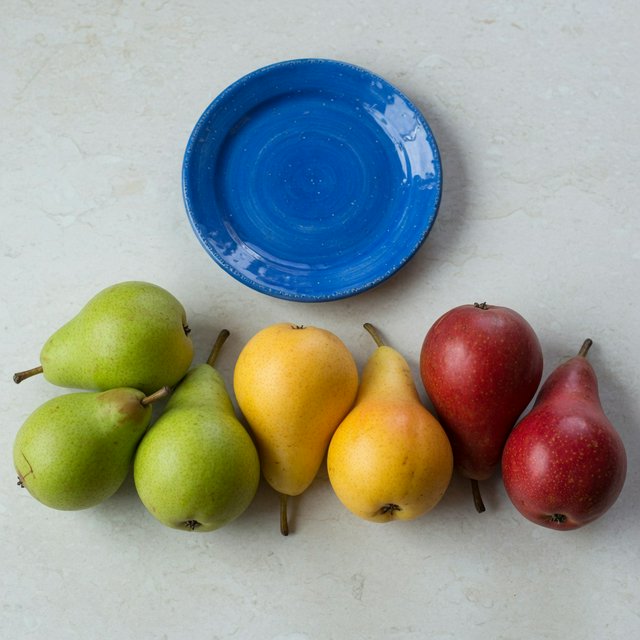 Exactly six ripe pears sit on a pale stone counter: two green pears on the left, two yellow pears in the center, and two red pears on the right. A small blue plate sits above the center pair, nothing else. — Grok Imagine Image Quality