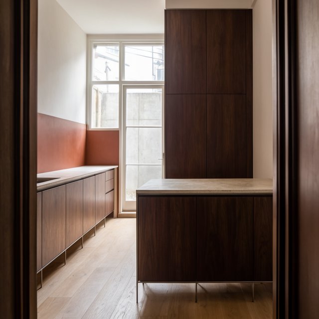 Along a galley kitchen, flat-front walnut cabinets meet pale limestone counters and a matte terracotta backsplash in restrained Scandinavian modern style; soft north-window daylight reveals the worktop from the doorway, with clean aligned lines and believable supports. — Model 11