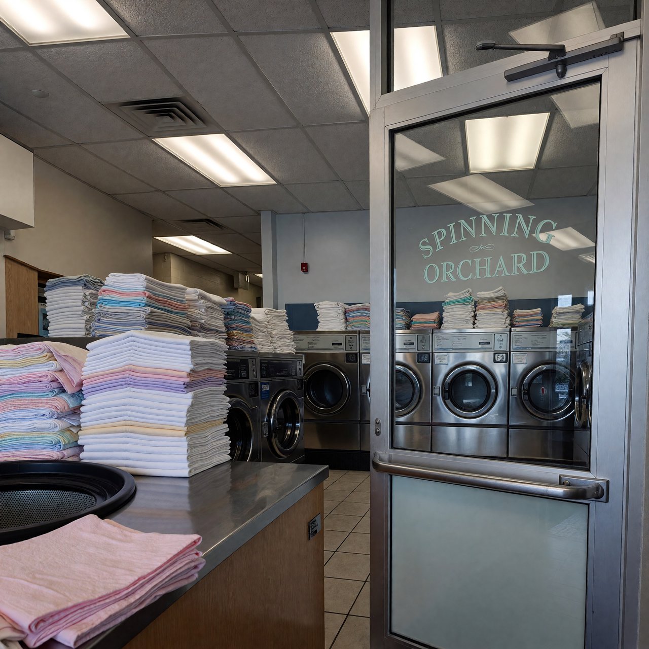 Inside a quiet neighborhood laundromat, chrome machines and folded linen glow beneath cool ceiling lights; "SPINNING ORCHARD" is printed in pale mint letters on the frosted glass entry door. — ImagineArt 1.5 Pro