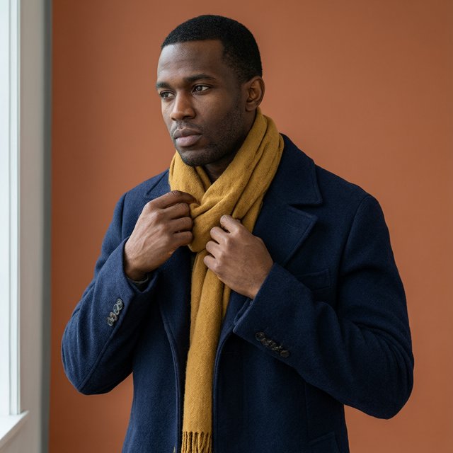 Editorial portrait of an adult Black man in his 30s wearing a deep navy wool overcoat and mustard scarf, fingers adjusting the scarf, thoughtful expression, muted terracotta studio backdrop, crisp cool window light. — Qwen Image 3.0 Pro