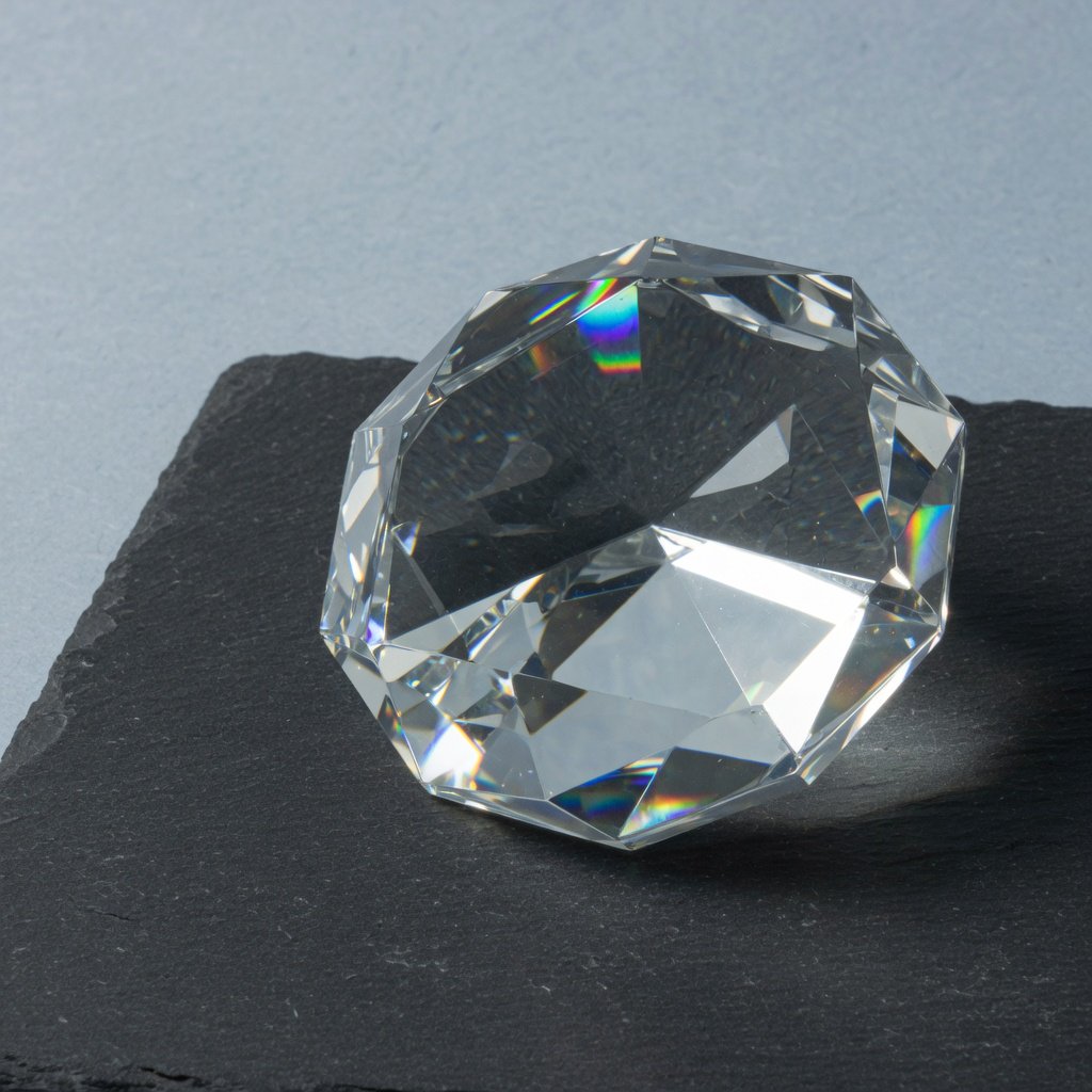 An unbranded, unmarked faceted optical-glass paperweight rests on charcoal slate against a pale blue-gray background; broad diffused studio light and a narrow side highlight reveal crisp internal refractions and bright edge reflections. — Grok Imagine Image Quality