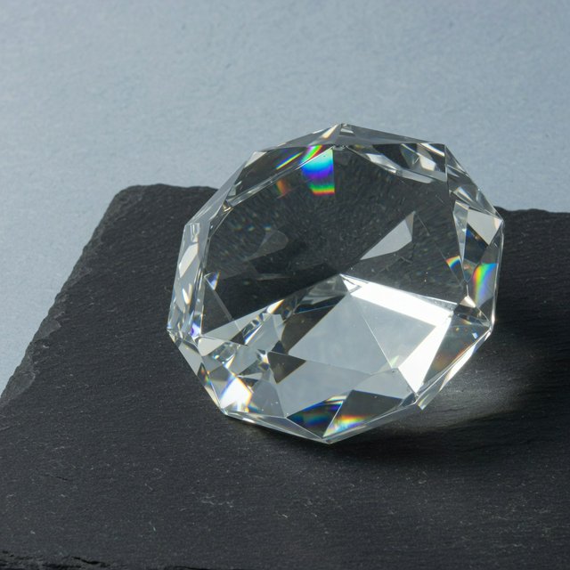 An unbranded, unmarked faceted optical-glass paperweight rests on charcoal slate against a pale blue-gray background; broad diffused studio light and a narrow side highlight reveal crisp internal refractions and bright edge reflections. — Grok Imagine Image Quality