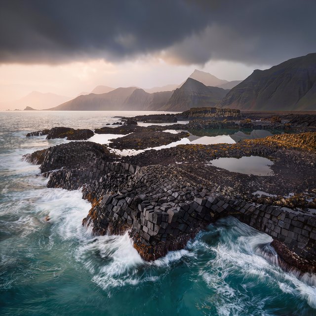A rugged volcanic coastline under clearing storm clouds at late afternoon, black basalt shelves descending into turquoise surf, sea spray clinging to jagged rocks, tide pools reflecting distant cliffs and layered mountains fading into rain haze. — Kling v3