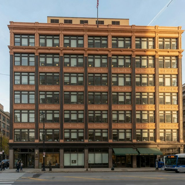 Exterior of a Chicago School office tower, with a visible steel-frame grid, warm terra-cotta cladding, broad rectangular windows, and a flat roofline; clear afternoon light, viewed from across a city street, with precise verticals and believable construction. — Grok Imagine Image Quality