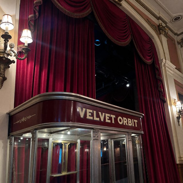 In a restored neighborhood cinema, brass sconces illuminate velvet curtains and a glass ticket booth; "VELVET ORBIT" is painted in ivory block letters across the booth’s burgundy fascia. — ImagineArt 1.5 Pro