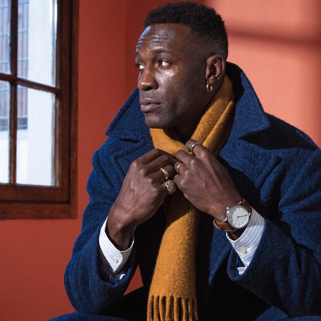 Editorial portrait of an adult Black man in his 30s wearing a deep navy wool overcoat and mustard scarf, fingers adjusting the scarf, thoughtful expression, muted terracotta studio backdrop, crisp cool window light. — Recraft v4