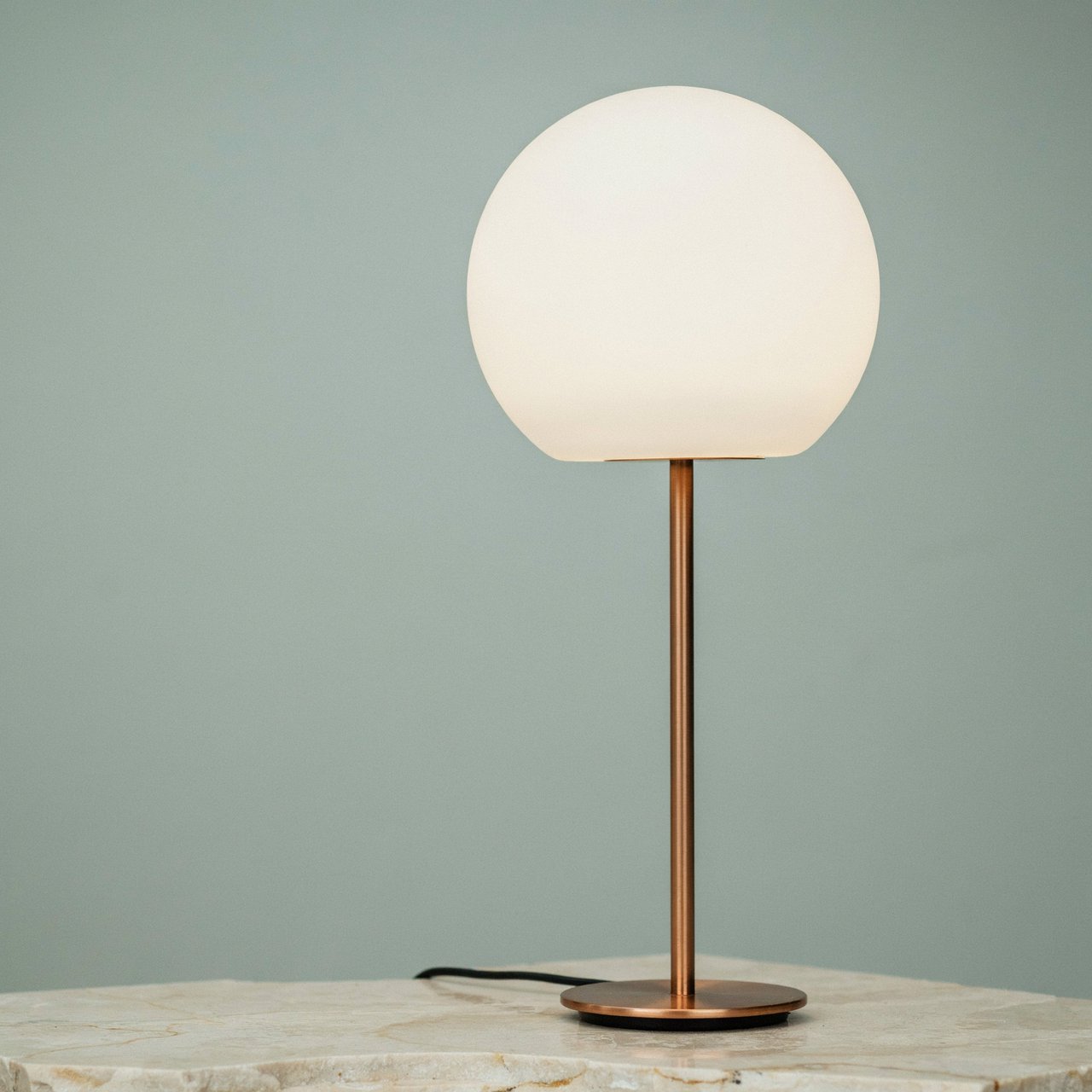 Commercial product photograph of an unbranded sculptural desk lamp with a frosted opal glass globe and brushed copper stem, standing on a pale travertine surface against a muted sage background, illuminated by soft overhead diffusion and a delicate warm rim light. — Ideogram v4.5