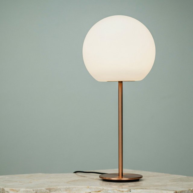 Commercial product photograph of an unbranded sculptural desk lamp with a frosted opal glass globe and brushed copper stem, standing on a pale travertine surface against a muted sage background, illuminated by soft overhead diffusion and a delicate warm rim light. — Ideogram v4.5
