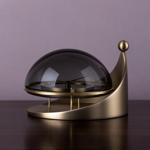 Commercial product photograph of an unbranded sculptural table clock made from satin brass and smoked glass, resting on a dark walnut surface against a muted lavender background, illuminated by a large diffused softbox from the upper left and a subtle rear rim light. — Krea 2 Large