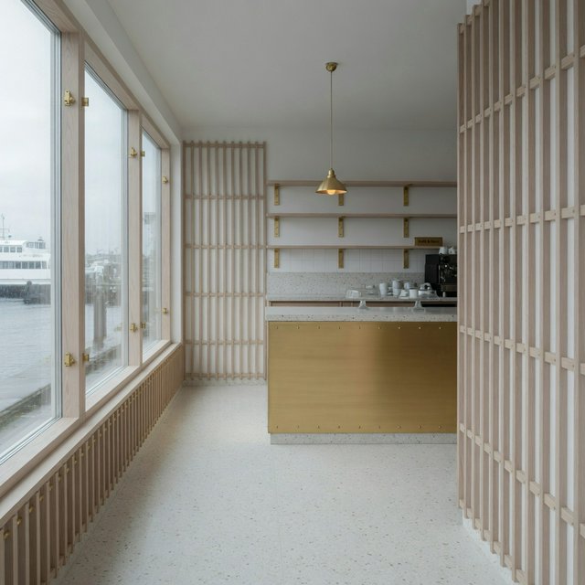 Inside an empty harbor café, restrained Danish modern design pairs ash slat screens, pale terrazzo, and brushed brass; overcast window light reaches the counter, viewed from the entrance, with aligned joinery and believable supports. — Model 6