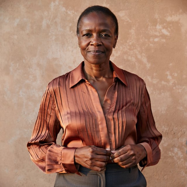Portrait of an adult Black woman in her 50s wearing a pleated copper satin blouse and tailored charcoal trousers, her hands gently holding the blouse fabric, calm half-smile, warm plaster backdrop, late-afternoon window light grazing her face. — ImagineArt 2