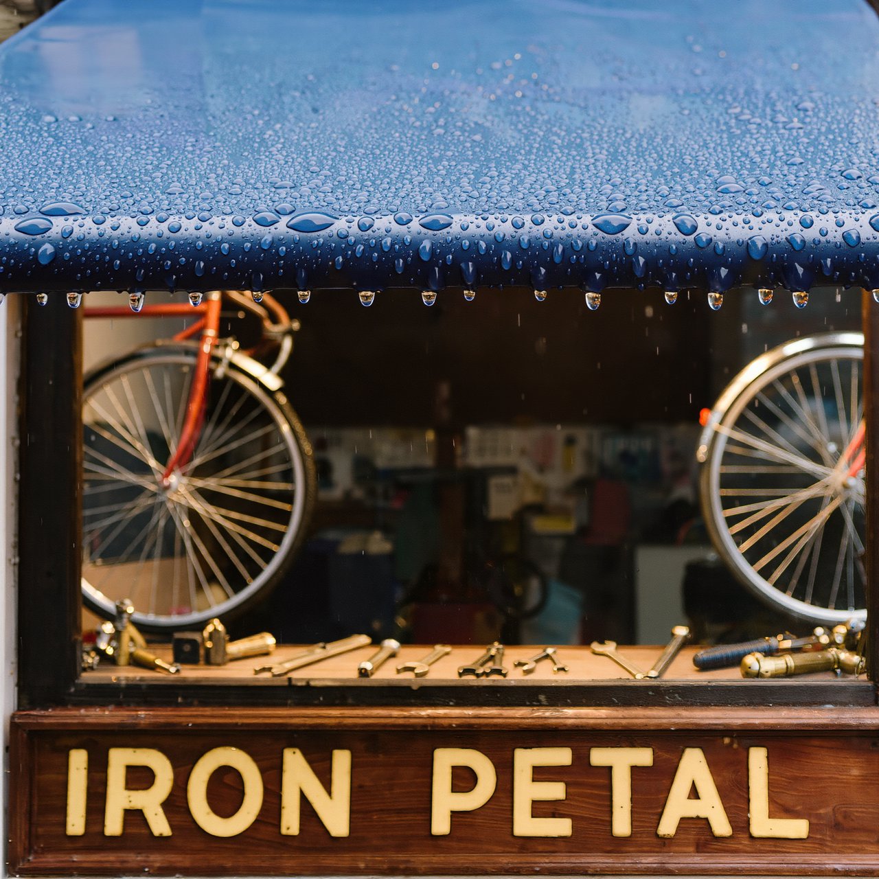 Rain beads on a bicycle repair shop’s blue awning, with wheels and brass tools visible behind the glass; “IRON PETAL” is lettered in warm yellow enamel capitals across the wooden fascia. — Kling v3