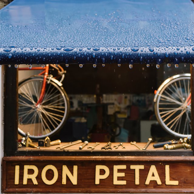 Rain beads on a bicycle repair shop’s blue awning, with wheels and brass tools visible behind the glass; “IRON PETAL” is lettered in warm yellow enamel capitals across the wooden fascia. — Kling v3