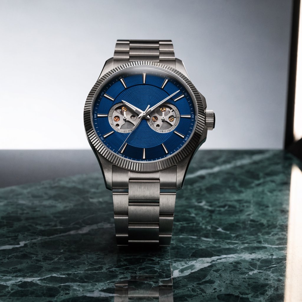 Commercial product photograph of an unbranded mechanical wristwatch with a brushed titanium case and deep blue enamel dial, resting on dark green marble against a soft gray background, illuminated by a large diffused softbox and narrow edge light. — Recraft v4