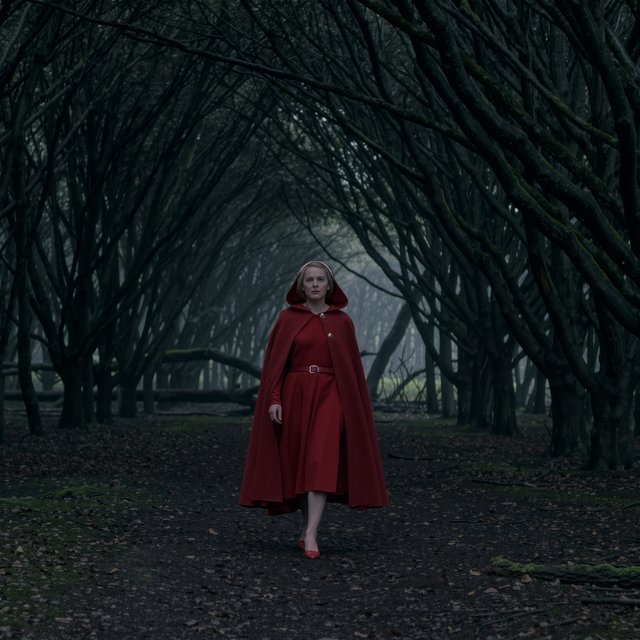 Medium shot at eye level: 1960s vintage film aesthetic. An adult woman with long, blonde hair walks alone through a dark forest. She wears a red hooded cloak, with the hood pulled up and fastened at the neck by a silver buckle. Photorealistic 1960s Technicolor film grain; soft, shallow depth of field. — Kling v3 Omni