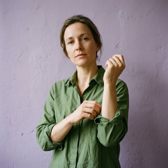 Portrait photograph of an adult woman in her late 30s wearing a moss-green cotton poplin shirt, fingers lightly turning its rolled cuff. Her calm, observant expression faces the camera against a chalky lavender backdrop, lit by soft diffused skylight. — Nano Banana 2.1