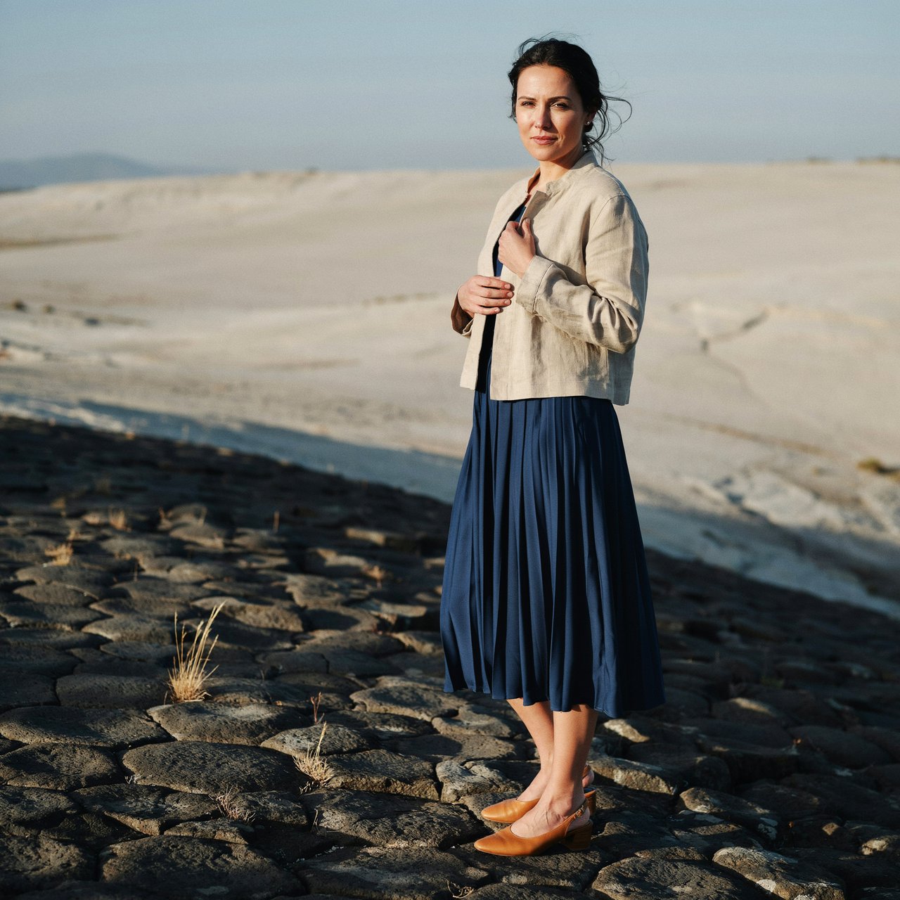 Across a basalt terrace, an adult woman in her early forties poses in a full-length fashion photograph, wearing a pleated indigo silk dress and cropped linen jacket, her composed half-smile lit by hard afternoon sunlight from one side against a pale limestone ridge. — Ideogram v4