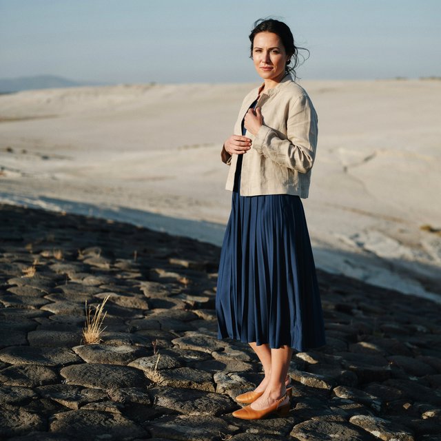 Across a basalt terrace, an adult woman in her early forties poses in a full-length fashion photograph, wearing a pleated indigo silk dress and cropped linen jacket, her composed half-smile lit by hard afternoon sunlight from one side against a pale limestone ridge. — Ideogram v4