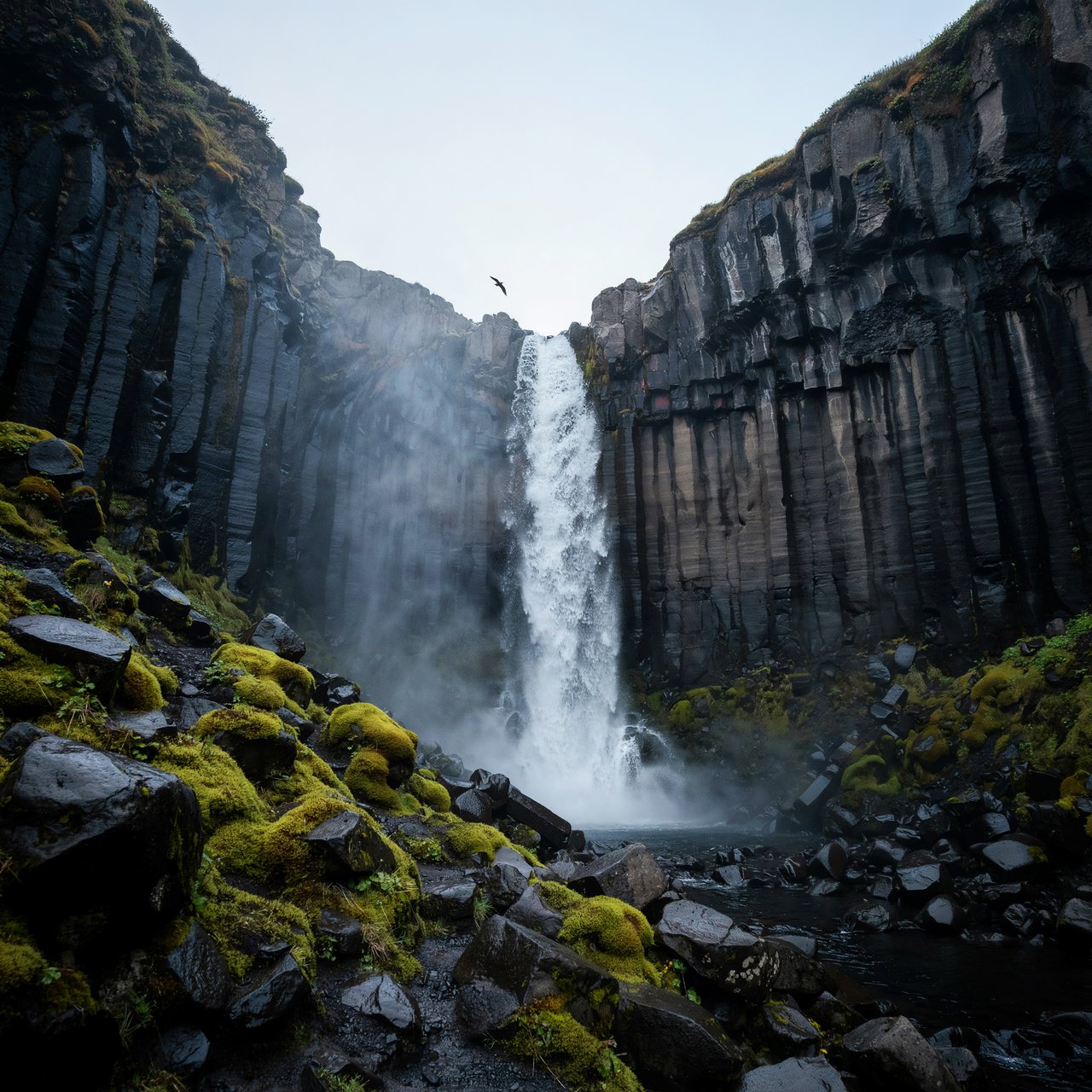 After a passing squall, a waterfall drops through a narrow basalt gorge at dusk, its white plume breaking against dark ledges; rain-beaded moss and sharp foreground stones lead toward cliffs fading behind drifting spray. — Ideogram v4